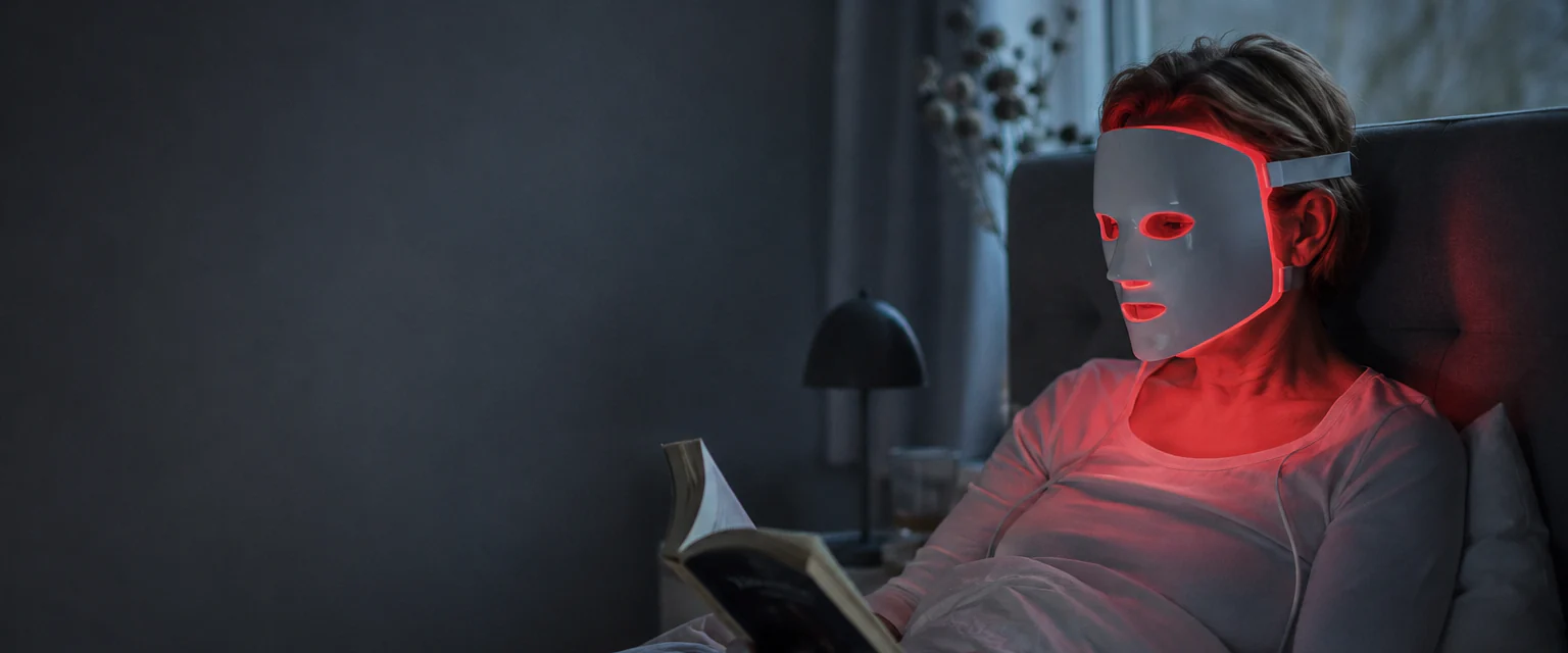 Kvinna läser en bok i sängen en kväll med en LED-mask som lyser rött